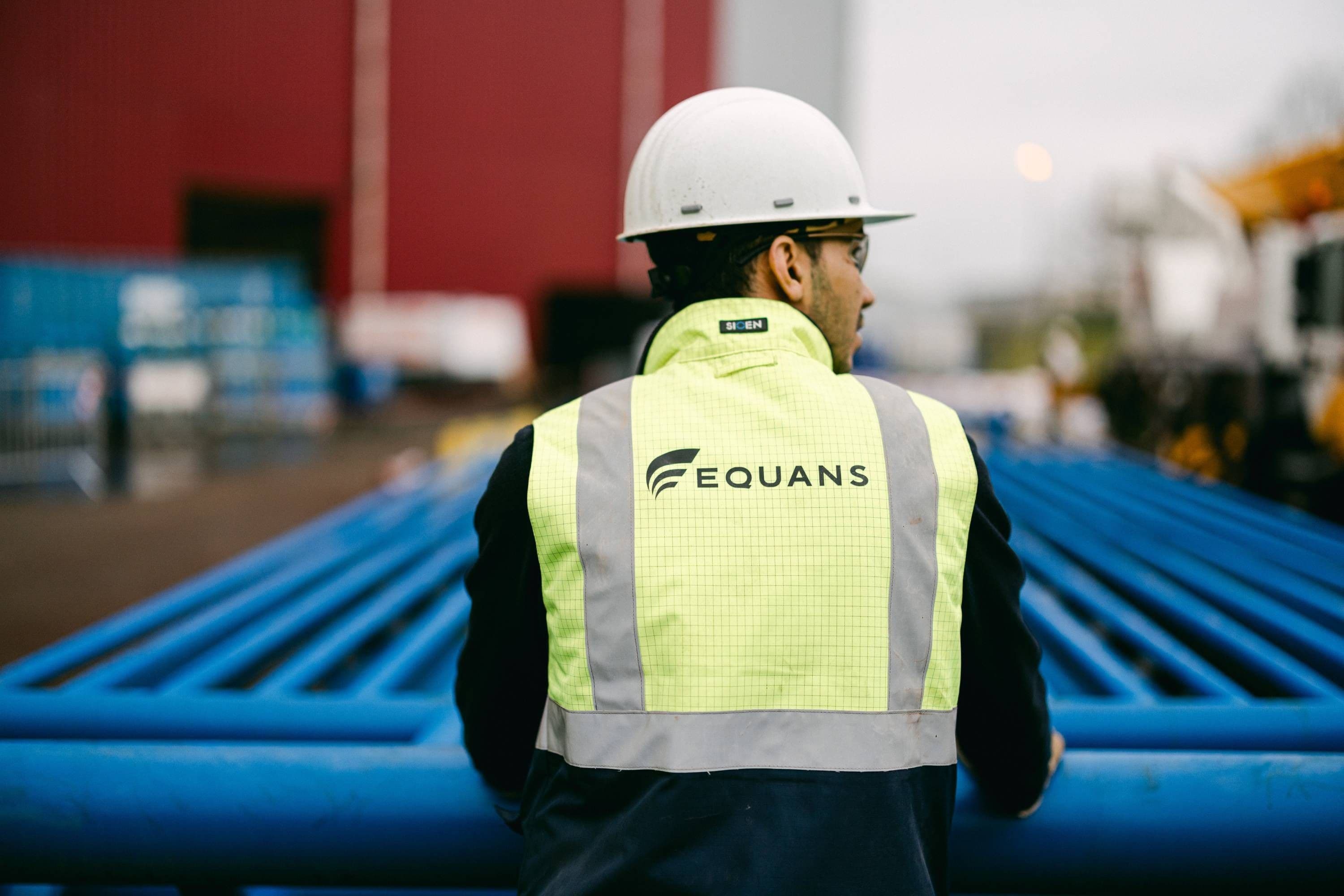 Equans engineer in a hard hat and hi-vis vest beside blue industrial pipework