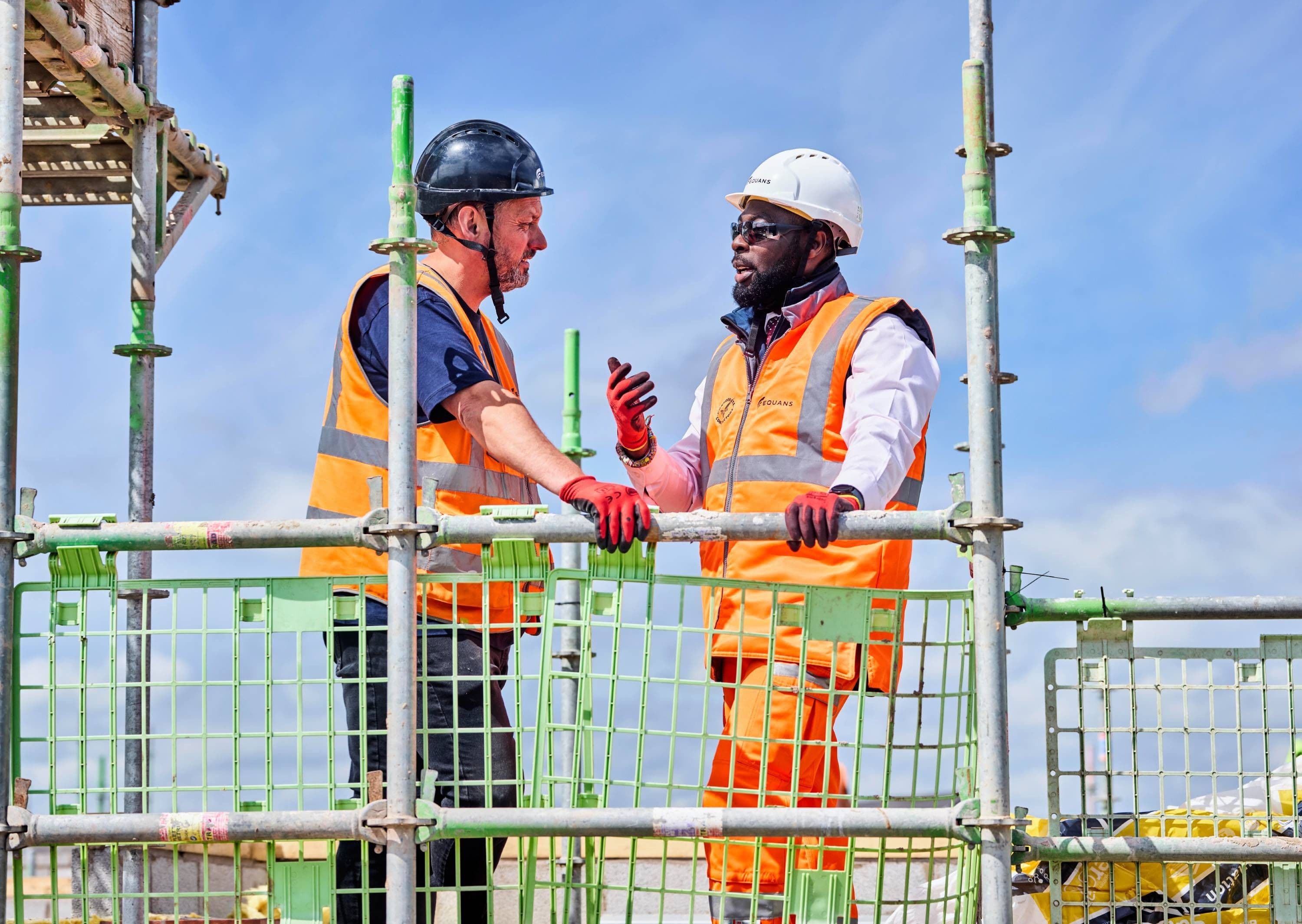 Two Equans workers in hi-vis on scaffolding against a blue sky